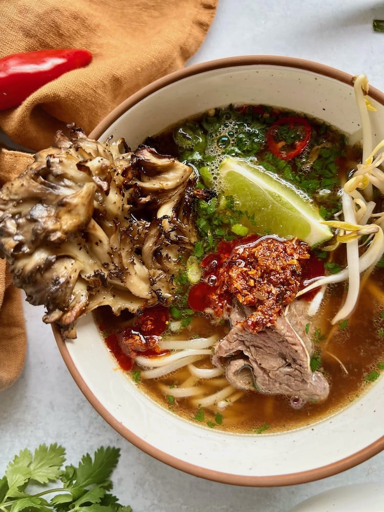 Beef Pho in a bowl with maitake mushroom, lime wedge, and noodles.