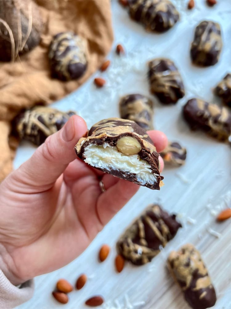 An Almond Coconut Candy Bar cut in half and held toward the viewer. Below diffused in the background are more candy bars