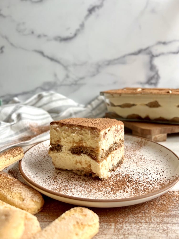 A slice of Guinness and Irish Cream Tiramisu on a plate dusted with cocoa powder with a pan of tiramisu in the background