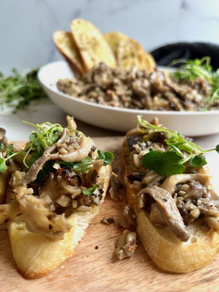 Mixed Mushroom Bruschetta on two crostini and in the background there's a serving dish filled with mushroom bruschetta and more crostini