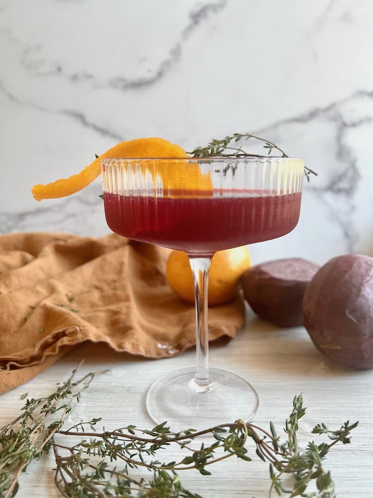 The Hangry Beet Cocktail in a coupe glass with orange peel and thyme sprig garnishes