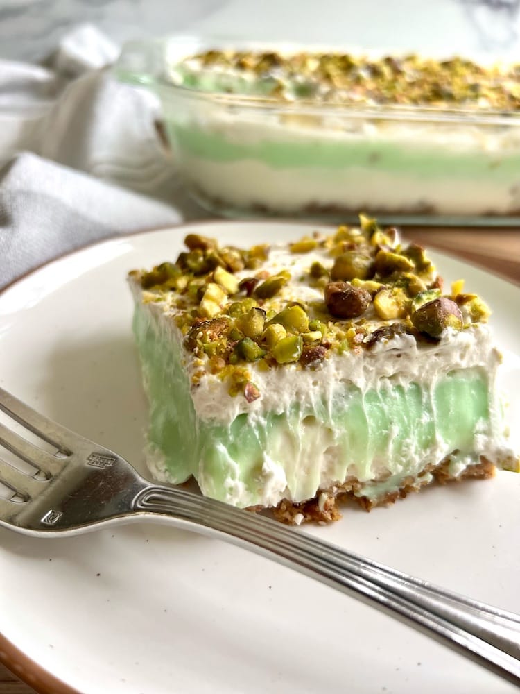 A slice of Grammie's Layered Pistachio Dessert on a plate with fork, and a pan of Grammie's Layered Pistachio Dessert in the background.