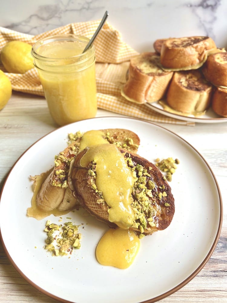Two slices of Pistachio French Toast with Lemon Curd on small plate. In the background is a mason jar of lemon curd and another plate of Pistachio French Toast