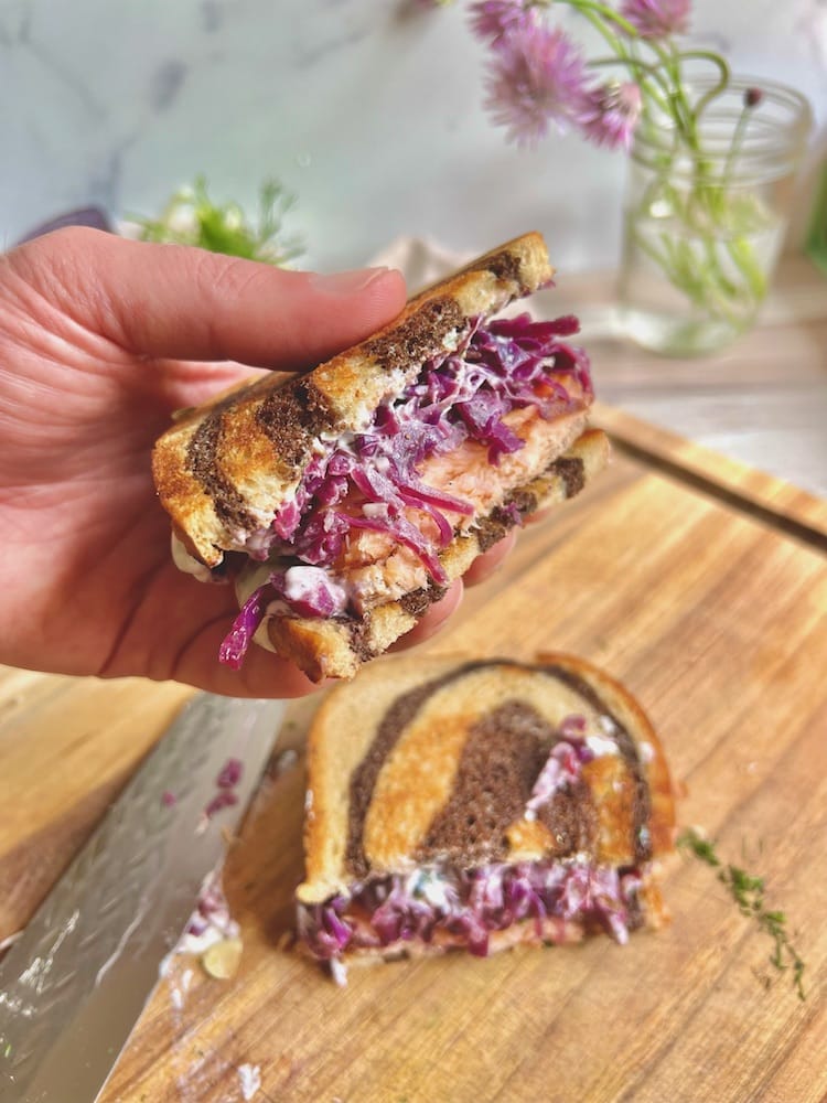 A Smoked Salmon Reuben cut in half with one half shown to the viewer above a cutting board and the other half of the sandwich