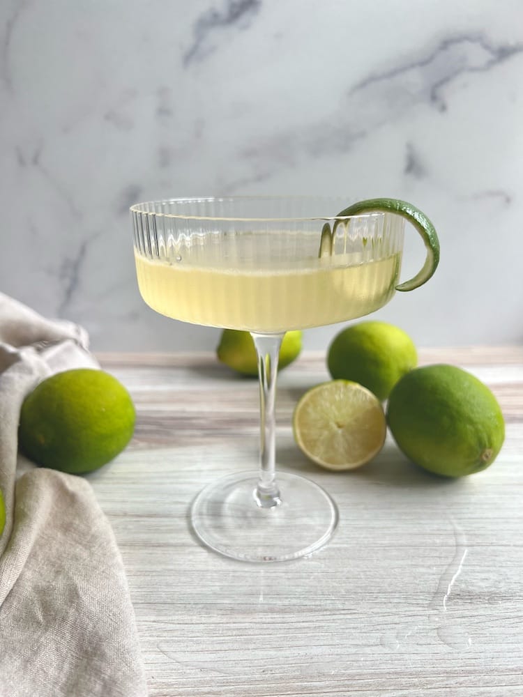 A Cruel Summer Cocktail in a coupe glass surrounded by fresh limes