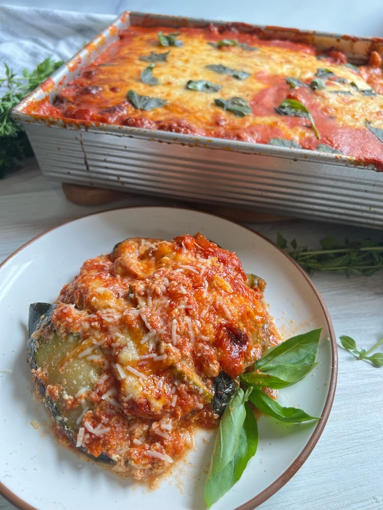A slice of Hangry Zucchini Lasagna on a plate next to a baking dish of lasagna