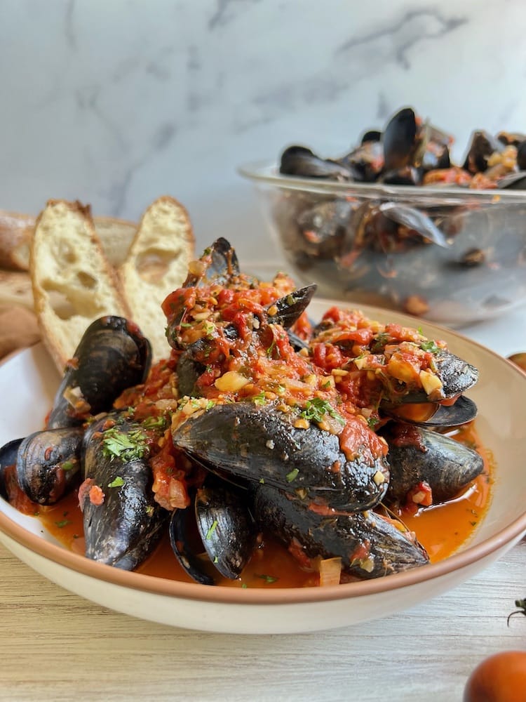 Italian Mussels in Tomato Broth in a bowl with two slices of bread and a larger clear bowl of mussels in the background