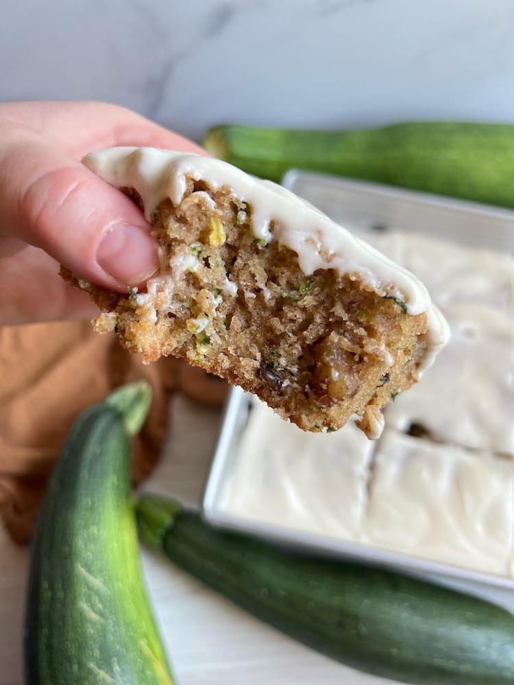 A slice of Zucchini Cake held toward the camera with one bite taken out of it. Below is the pan of Zucchini Cake