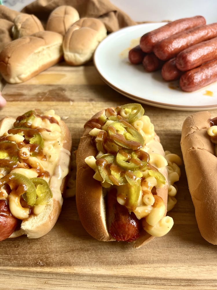 Mac N Cheese Hot Dogs on a cutting board with a plate of hot dogs and buns behind them