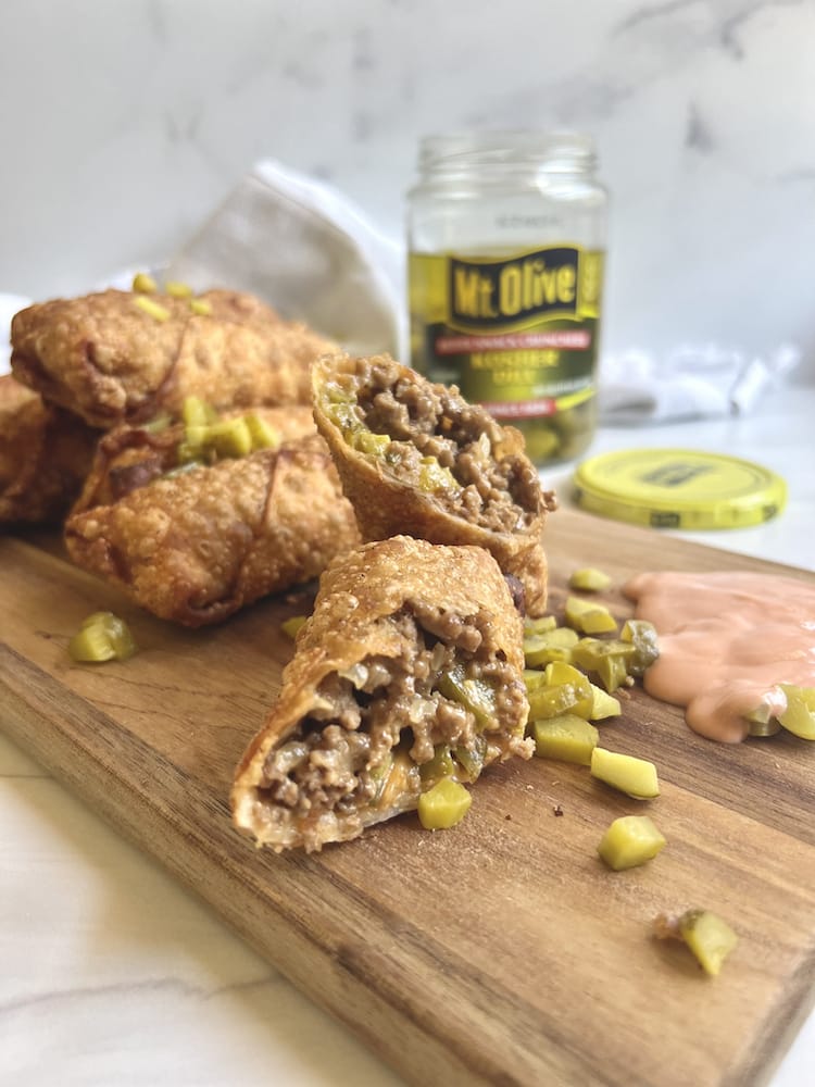 Cheeseburger Egg Rolls on a cutting board with a jar of Mt. Olive Pickles in the background