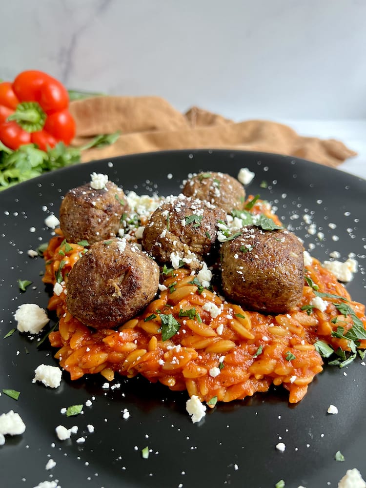 Lamb Meatballs with Roasted Pepper Orzo on a black plate with red bell peppers in the background