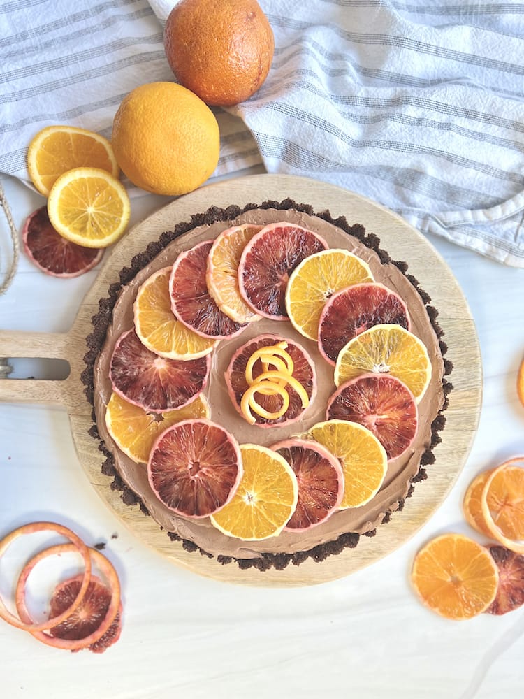 A Chocolate Orange Tart on a cutting board surrounded with fresh oranges, some sliced and some unsliced