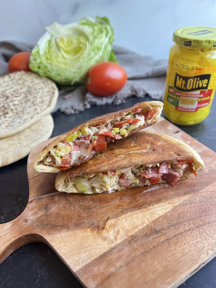 Italian Hoagie Dip Pitas on a cutting board with Mt Olive Banana Peppers, fresh lettuce, tomato, and more pitas in the background
