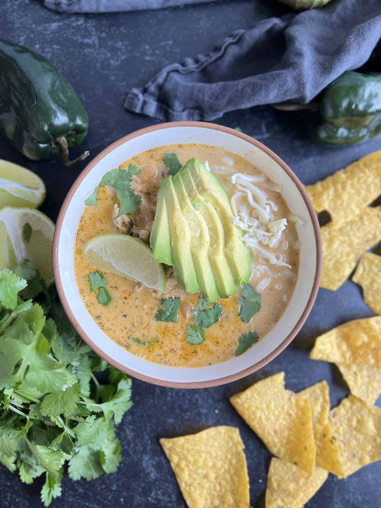 White Chicken Chili in a bowl garnished with sliced avocado, Monterey Jack cheese, lime wedge, and fresh cilantro. Bowl is surrounded by tortilla chips and more fresh cilantro