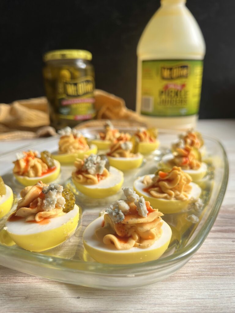 Buffalo Deviled Eggs on a glass serving dish with a jar of pickles and Mt. Olive Pickle Juicers in the background