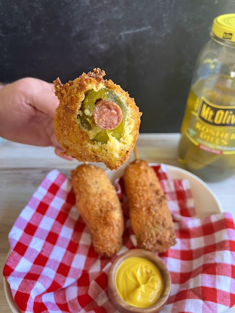 A Crunchy Pickle Corn Dog with a bite taken out of the top of it to show the hot dog and pickle inside.