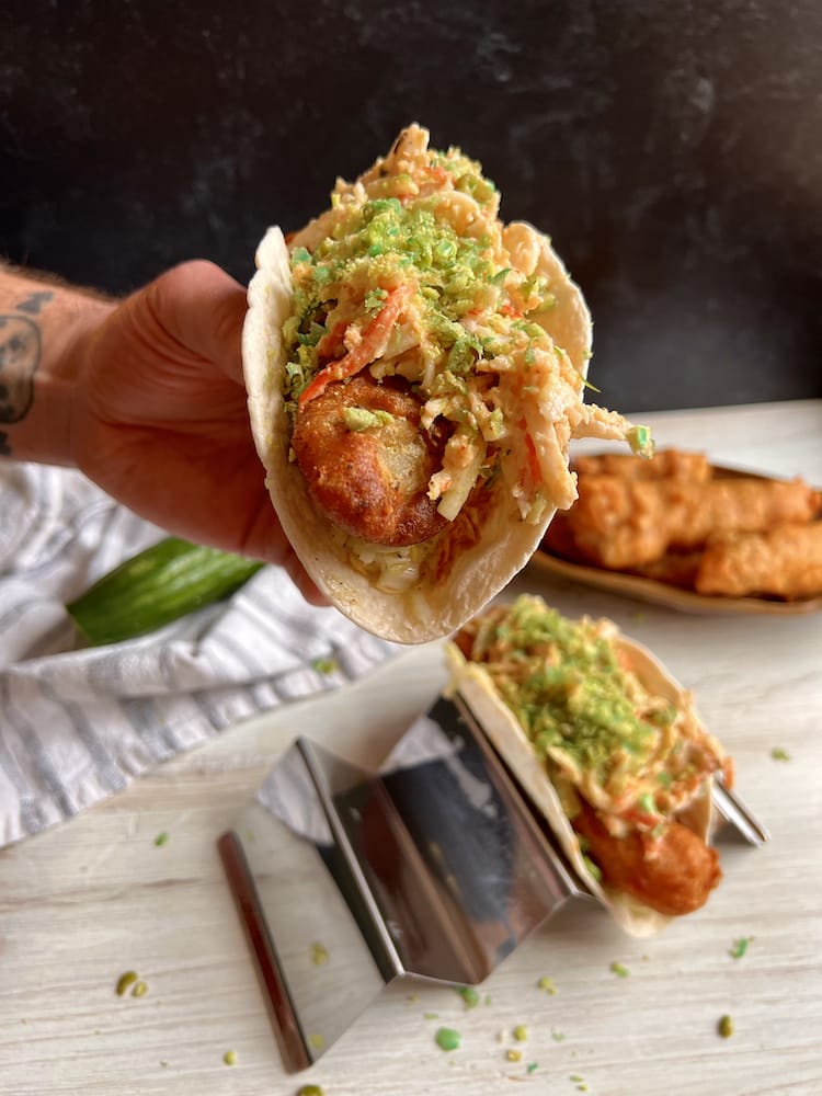 A Beer Battered Fish Taco held in one hand toward the viewer with another Beer Battered Fish Taco below in a taco rack