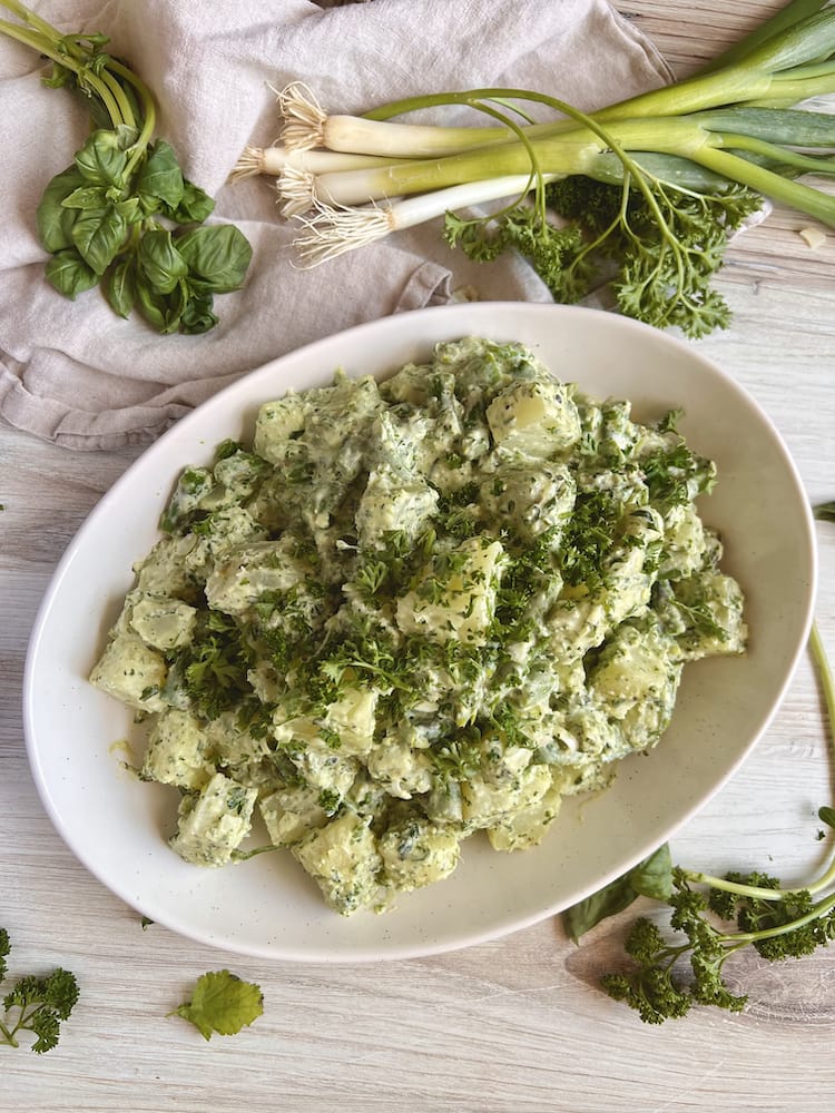 Green Goddess Potato Salad in a white serving dish with fresh scallions and herbs surrounding it