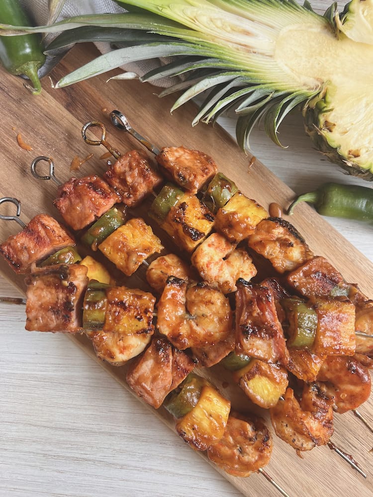 Hawaiian Ham and Shrimp Kabobs on a wooden cutting board with a fresh pineapple sliced in half next to them