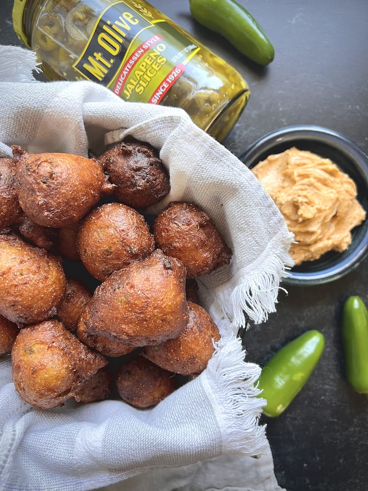 Jalapeño Hush Puppies in a linen-lined basket next to a small bowl of spiced honey butter and a jar of Mt. Olive Jalapeños