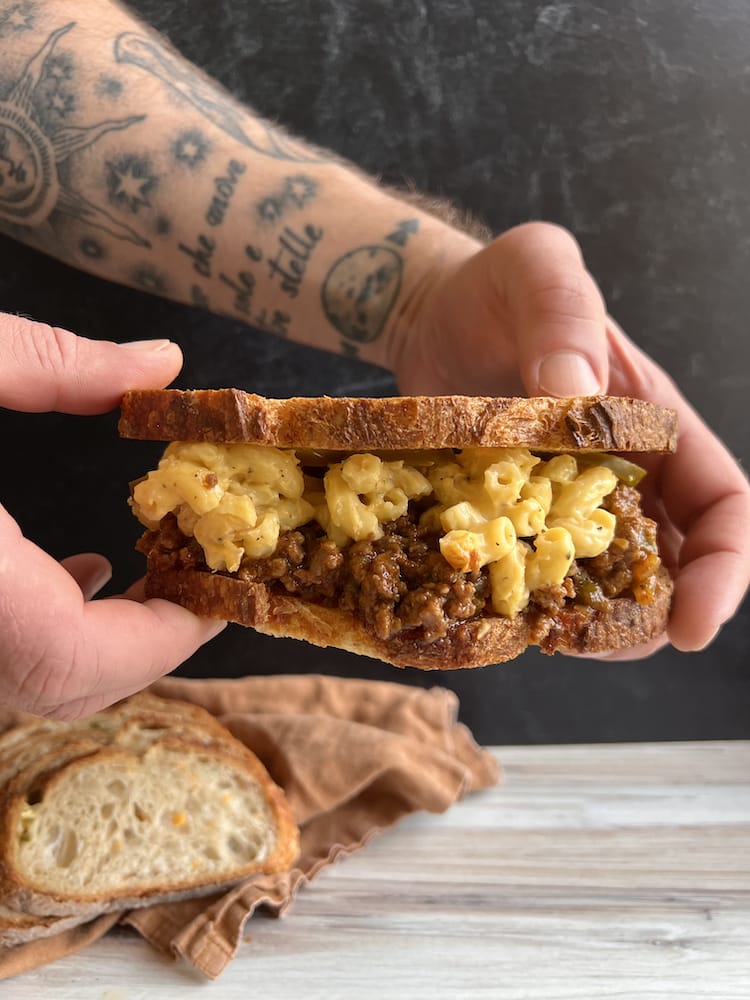 A Sloppy Joe Mac and Cheese Sandwich held by two hands with fresh sliced bread in the background