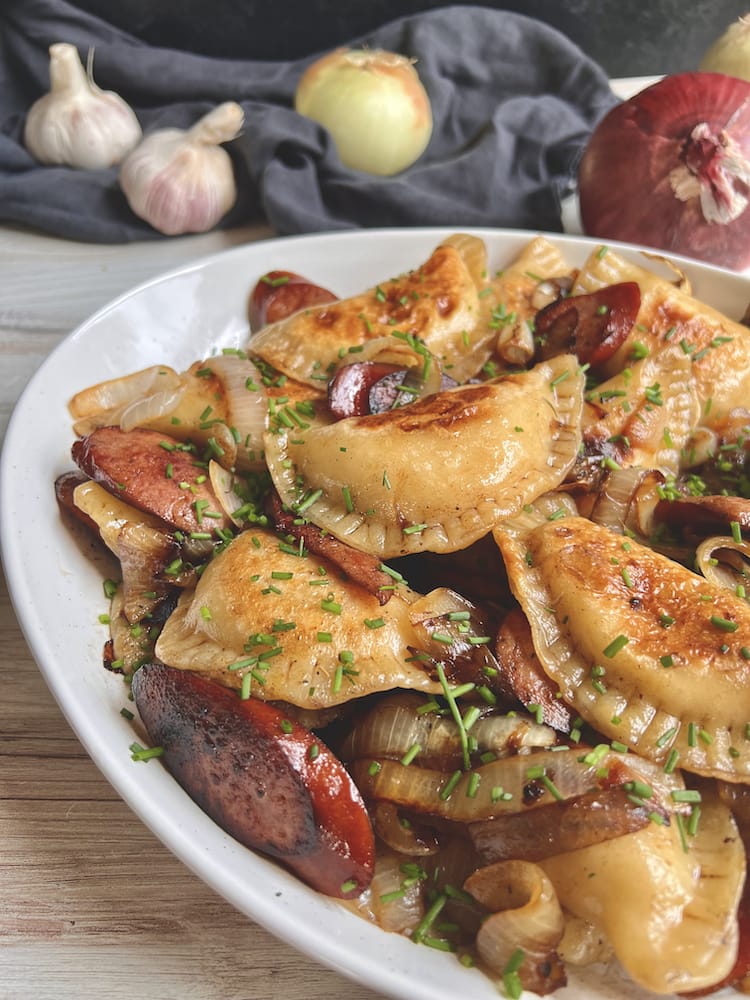 Griddled Pierogi and Kielbasa in a large white serving dish with fresh garlic and onion in the background