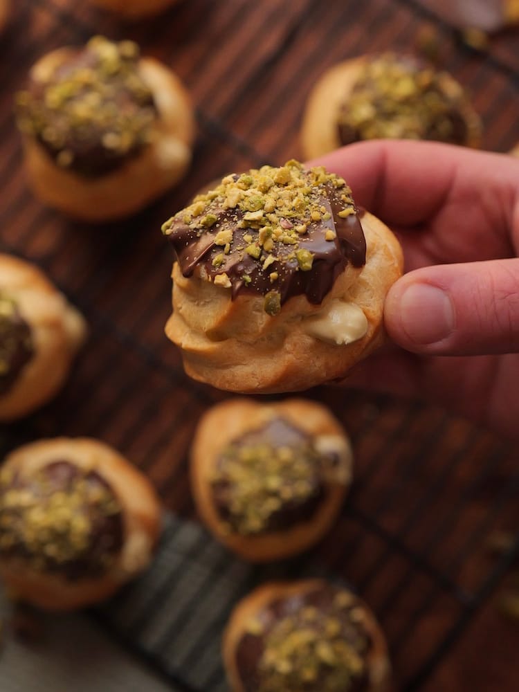 Pistachio Cream Puffs on a wire cooling rack with one held by a hand closest to the camera