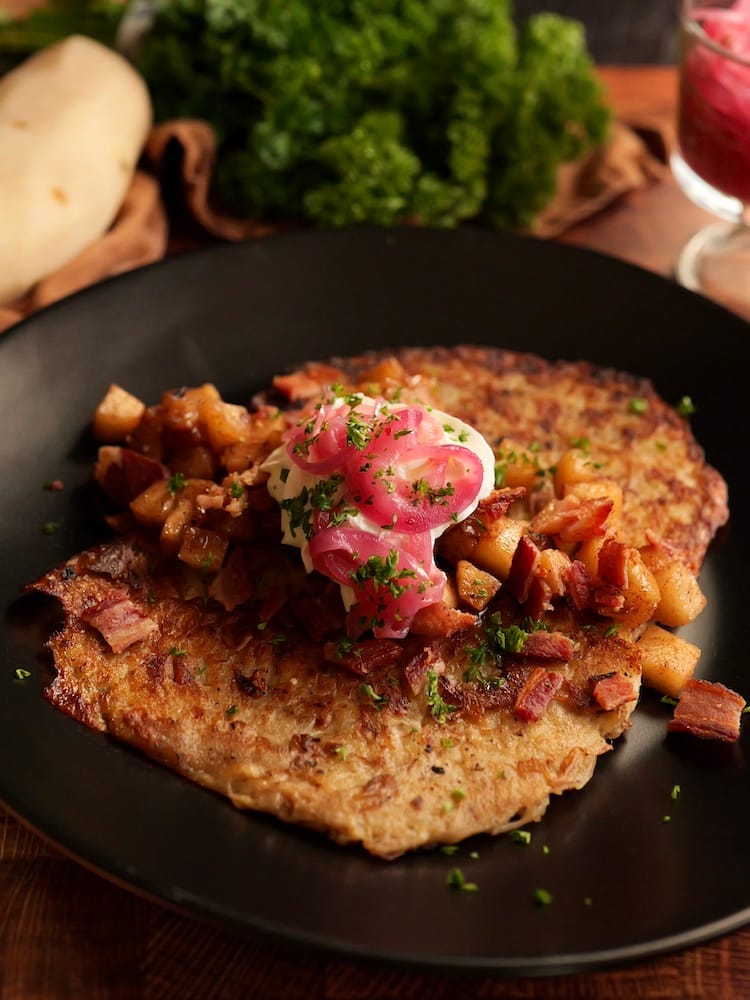 Sweet and Salty Latkes topped with pickled red onion, caramelized apples, bacon, and sour cream on a black plate