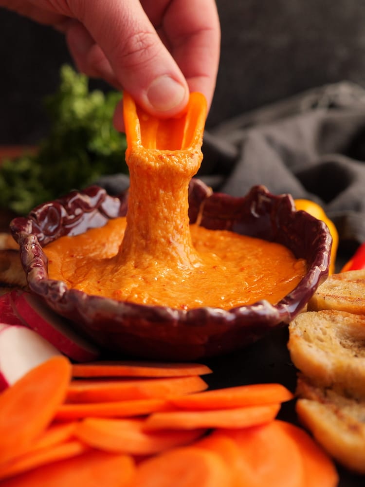 Fonduta di 'Nduja in a small dish with a sweet pepper piece being dipped by a hand into it so that there's a cheese pull from the fonduta up to the pepper