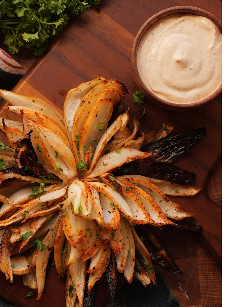 A Grilled Blooming Onion on a cutting board with a ramekin of dipping sauce next to it