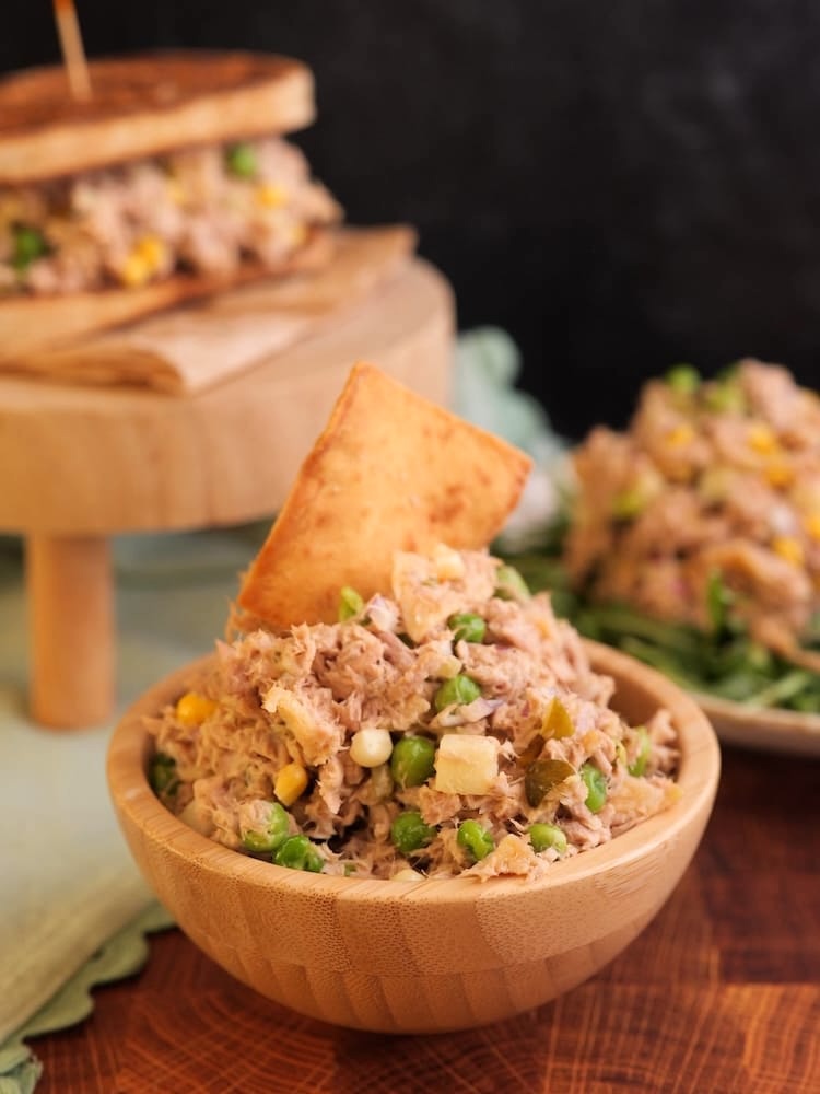 Matthew McConaughey Tuna Salad in a wooden bowl with a pita chip sticking out the top of the salad and in the background diffused are a tuna salad sandwich and tuna salad on arugula