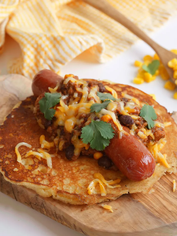 Corn Cake Chili Dog on wooden cutting board with wooden spoon and yellow staging cloth in background
