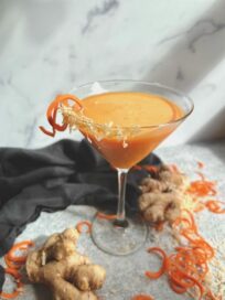 Ginger Carrot Juice Cocktail in Martini Glass surrounded by fresh ginger and carrots