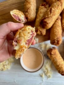 Half of a Reuben Egg Roll held up to the camera to show the sauerkraut and corned beef inside. More Reuben Egg Rolls are on a cutting board below.