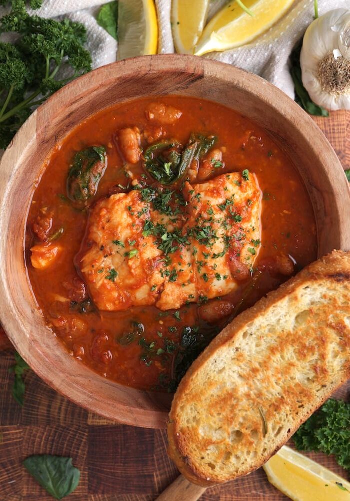 Tuscan Fish Stew in a wooden bowl with a slice of toasted bread on the rim of the bowl and the bowl surrounded by lemon wedges, fresh parsley, and a head of garlic